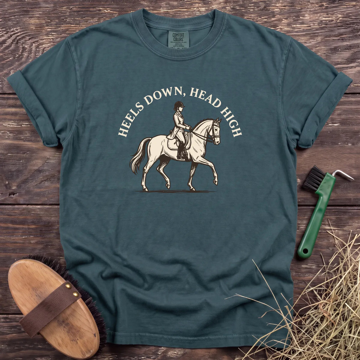 Teal t-shirt featuring a rider on a horse with the phrase "Heels Down, Head High" in a stylish design.