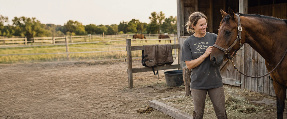 A rider in a Wild Rein Club graphic tee standing with her bay horse outside the barn.