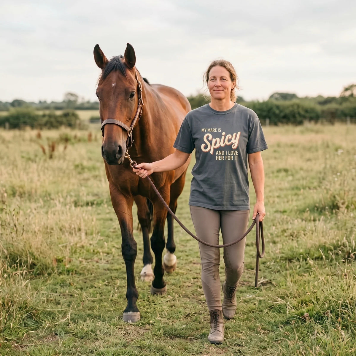 A rider wearing a My Mare Is Spicy tee walking beside her horse through a meadow.
